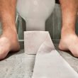 A roll of toilet paper at the feet of someone sitting on a toilet in a bathroom.