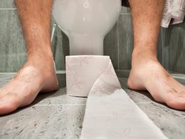 A roll of toilet paper at the feet of someone sitting on a toilet in a bathroom.