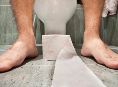 A roll of toilet paper at the feet of someone sitting on a toilet in a bathroom.