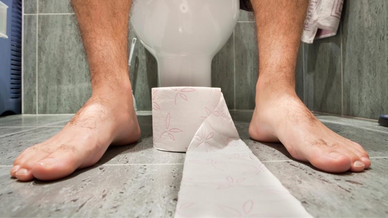 A roll of toilet paper at the feet of someone sitting on a toilet in a bathroom.