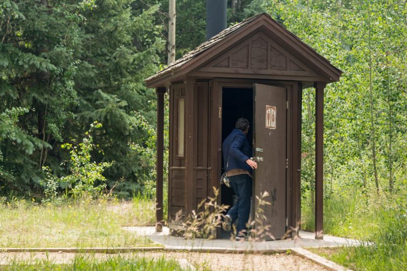 A person entering an outdoor restroom with a vault toilet at a park.