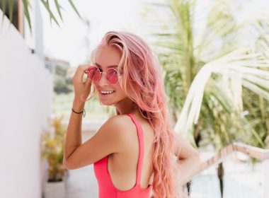 A woman with pink hair, sunglasses, and bathing suit smiles as she walks through a tropical resort.