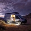 A Class B van parked in the desert at dusk and illuminated with electric lights inside and outside.