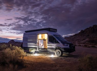A Class B van parked in the desert at dusk and illuminated with electric lights inside and outside.