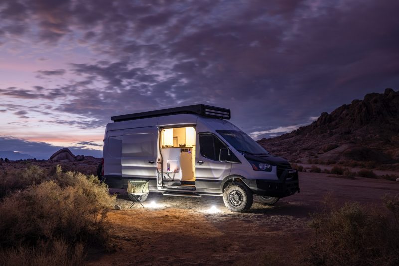 A Class B van parked in the desert at dusk and illuminated with electric lights inside and outside.