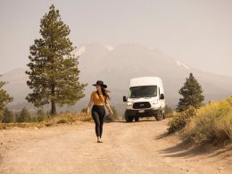 A woman in leggins and a hat walks away from her broken down van in the wilderness.