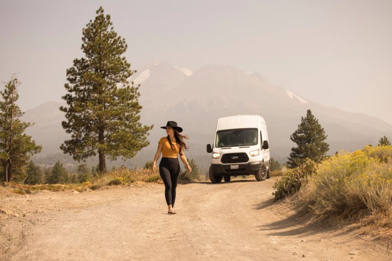 A woman in leggins and a hat walks away from her broken down van in the wilderness.