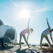 Two young campers do yoga on the beach outside of their teardrop travel trailer.