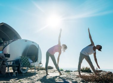 Two young campers do yoga on the beach outside of their teardrop travel trailer.