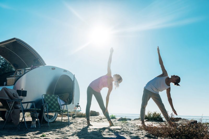 Two young campers do yoga on the beach outside of their teardrop travel trailer.