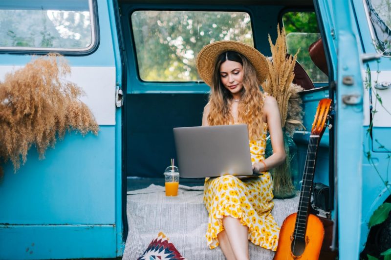 A beautiful woman with long hair and a dress works on her computer from her camper van and lives the van life on the road.