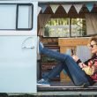A woman plays on her phone while sitting in the doorway of her campervan.