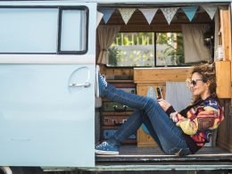 A woman plays on her phone while sitting in the doorway of her campervan.