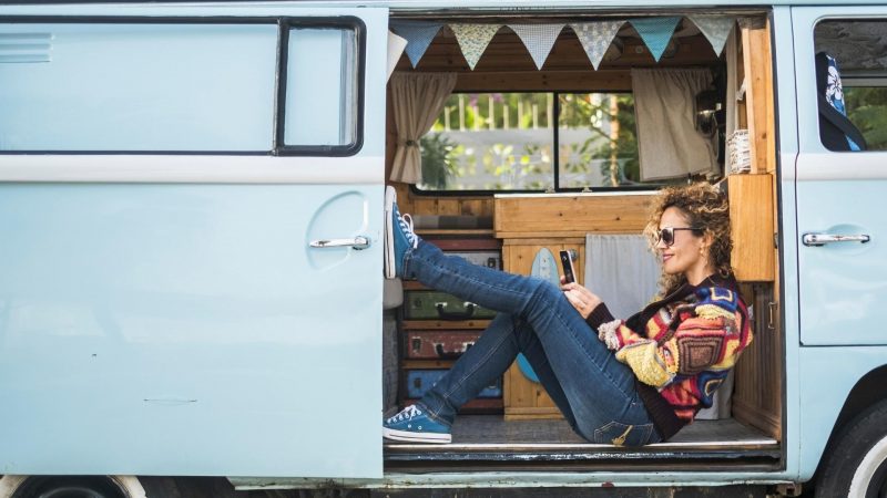 A woman plays on her phone while sitting in the doorway of her campervan.