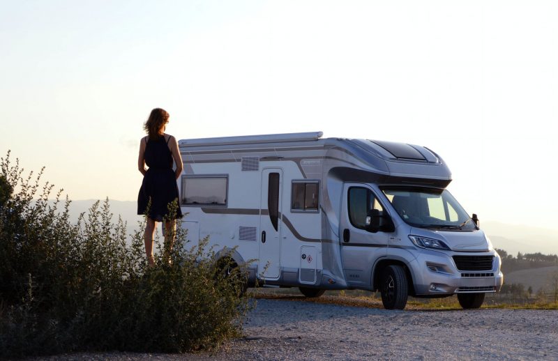 Silhouette of a woman standing outside of a class c motorhome as the sun sets over a field.