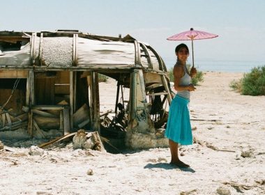 A woman with an umbrella stands beside a deteriorating RV in the desert sand.