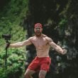 A muscley shirtless man in the forest yells and holds his video camera