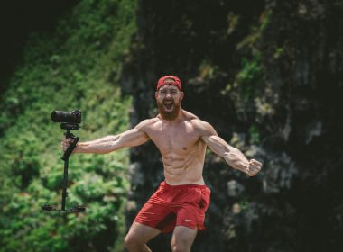 A muscley shirtless man in the forest yells and holds his video camera