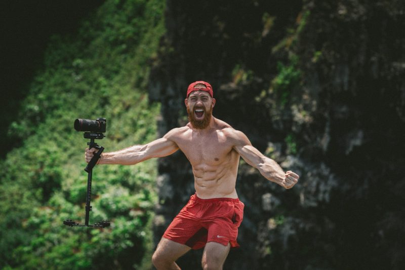 A muscley shirtless man in the forest yells and holds his video camera