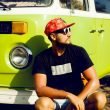 A cool guy with a hat and sunglasses squats down in front of his lime green vintage camper van.