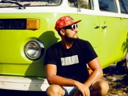 A cool guy with a hat and sunglasses squats down in front of his lime green vintage camper van.