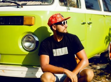 A cool guy with a hat and sunglasses squats down in front of his lime green vintage camper van.