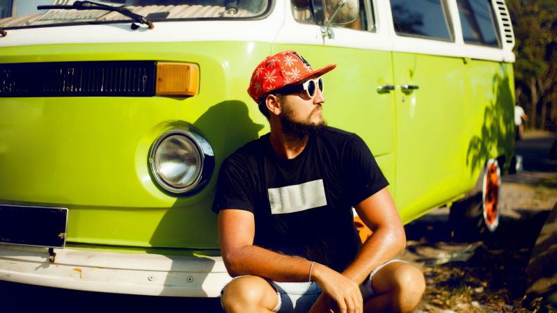 A cool guy with a hat and sunglasses squats down in front of his lime green vintage camper van.