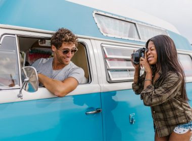 A woman photographs a man in the drivers seat of a van.