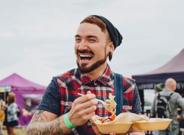 A man with a beanie, big earring, and a plaid shirt smiles and enjoys street food at a festival.