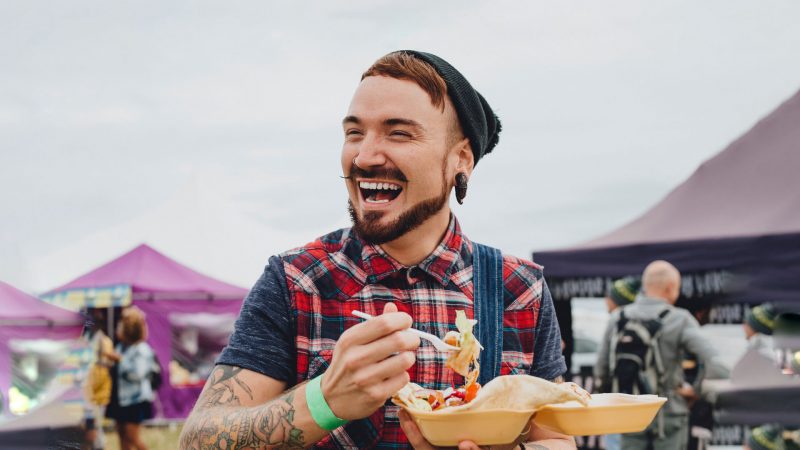A man with a beanie, big earring, and a plaid shirt smiles and enjoys street food at a festival.