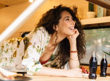 A beautiful woman with curly brunette hair smiles and looks out her travel trailer window.