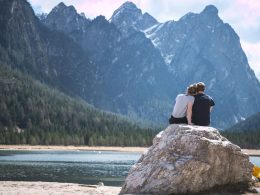 A traveling couple sits on a boulder overlooking a lake and mountains.