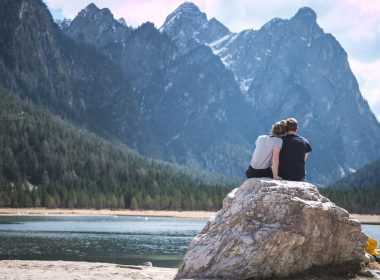 A traveling couple sits on a boulder overlooking a lake and mountains.