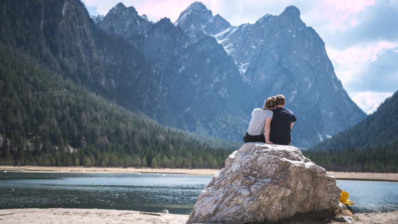 A traveling couple sits on a boulder overlooking a lake and mountains.
