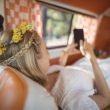 A boho woman with flowers in her hair lays in her camper van and works on her phone.
