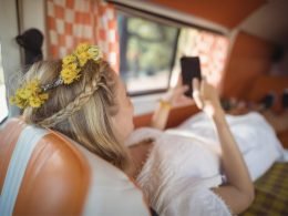 A boho woman with flowers in her hair lays in her camper van and works on her phone.