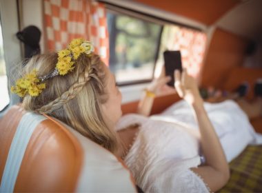 A boho woman with flowers in her hair lays in her camper van and works on her phone.