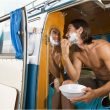 A shirtless man shaves using a mirror in his camper van