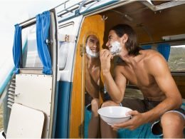 A shirtless man shaves using a mirror in his camper van