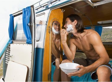 A shirtless man shaves using a mirror in his camper van