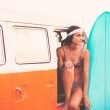 A woman sits in the open door of her camper van holding her surf board.