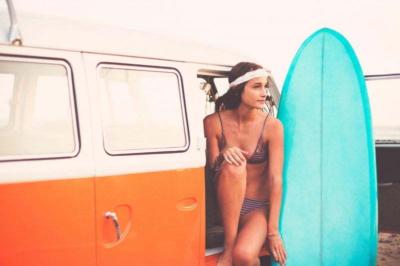 A woman sits in the open door of her camper van holding her surf board.