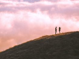 A couple of adventurers stand high up in a mountain with clouds all around them.