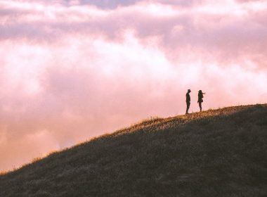 A couple of adventurers stand high up in a mountain with clouds all around them.