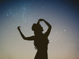A silhoutte of a woman with long curly hair and hat against a starry dusk sky.