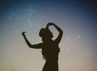 A silhoutte of a woman with long curly hair and hat against a starry dusk sky.