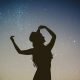 A silhoutte of a woman with long curly hair and hat against a starry dusk sky.