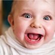 Up close of a goofy baby face with eyes wide and mouth agape showing two little teeth.