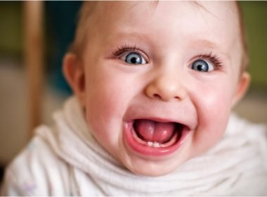 Up close of a goofy baby face with eyes wide and mouth agape showing two little teeth.