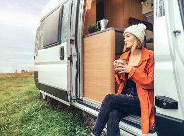 A woman sits out the doorway in her camper van drinking coffee in a valley.